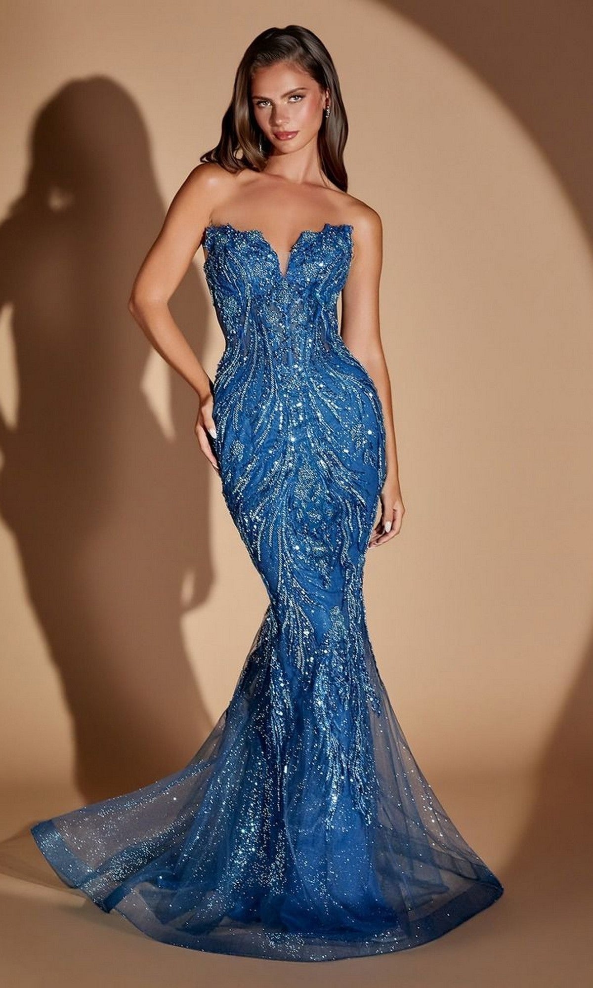 A woman wearing the Long Prom Dress: Ladivine CM393—a strapless, form-fitting blue gown adorned with shimmering sequin details and a flared sheer train—stands against a beige background.